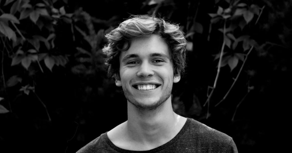 A black and white portrait of a young man smiling outdoors with natural background.